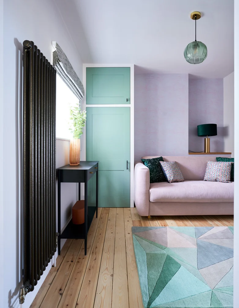 Home transformation a cosy and colourful living space with lilac walls and sage green built in cabinets, finished off with a blush pink sofa and geometric rug, the space has a hanging pendant light and a teal green lamp.