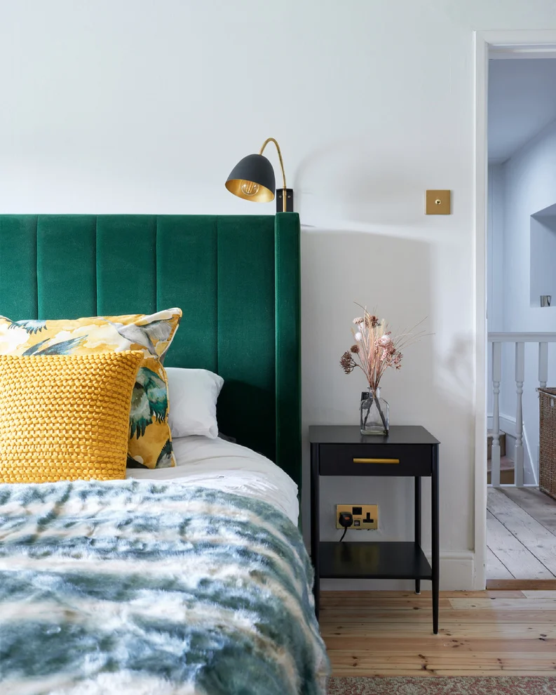 Home transformation this stylish bedroom has been completed with an emerald green velvet headboard and bright cushions with a patterned throw - the space is completed with a black and gold wall lamp for reading.