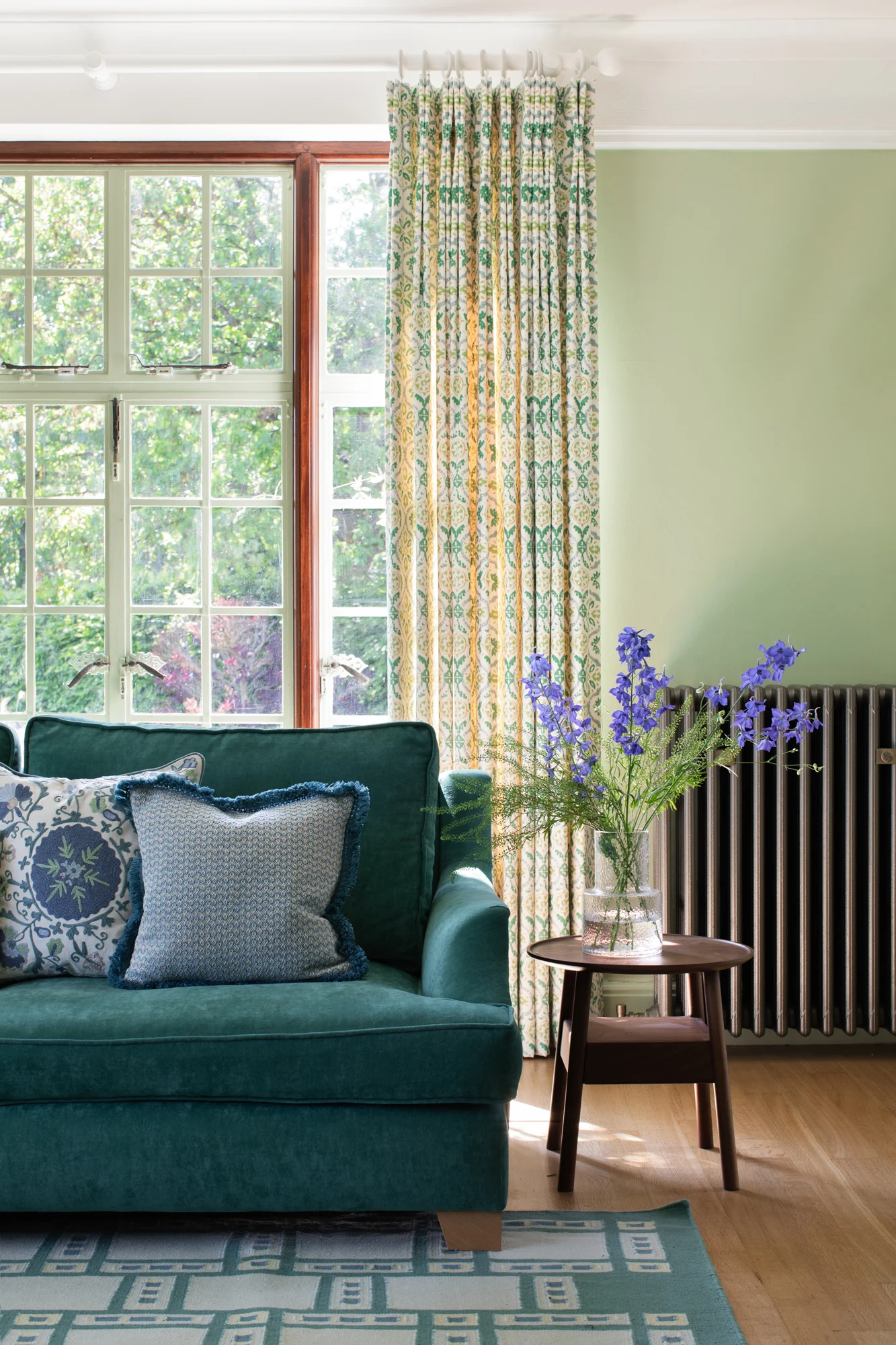 This lounge boasts garden-inspired interior design in a light-filled space with a teal sofa and floral cushions, finished with patterned curtains, and views of the blooming garden.