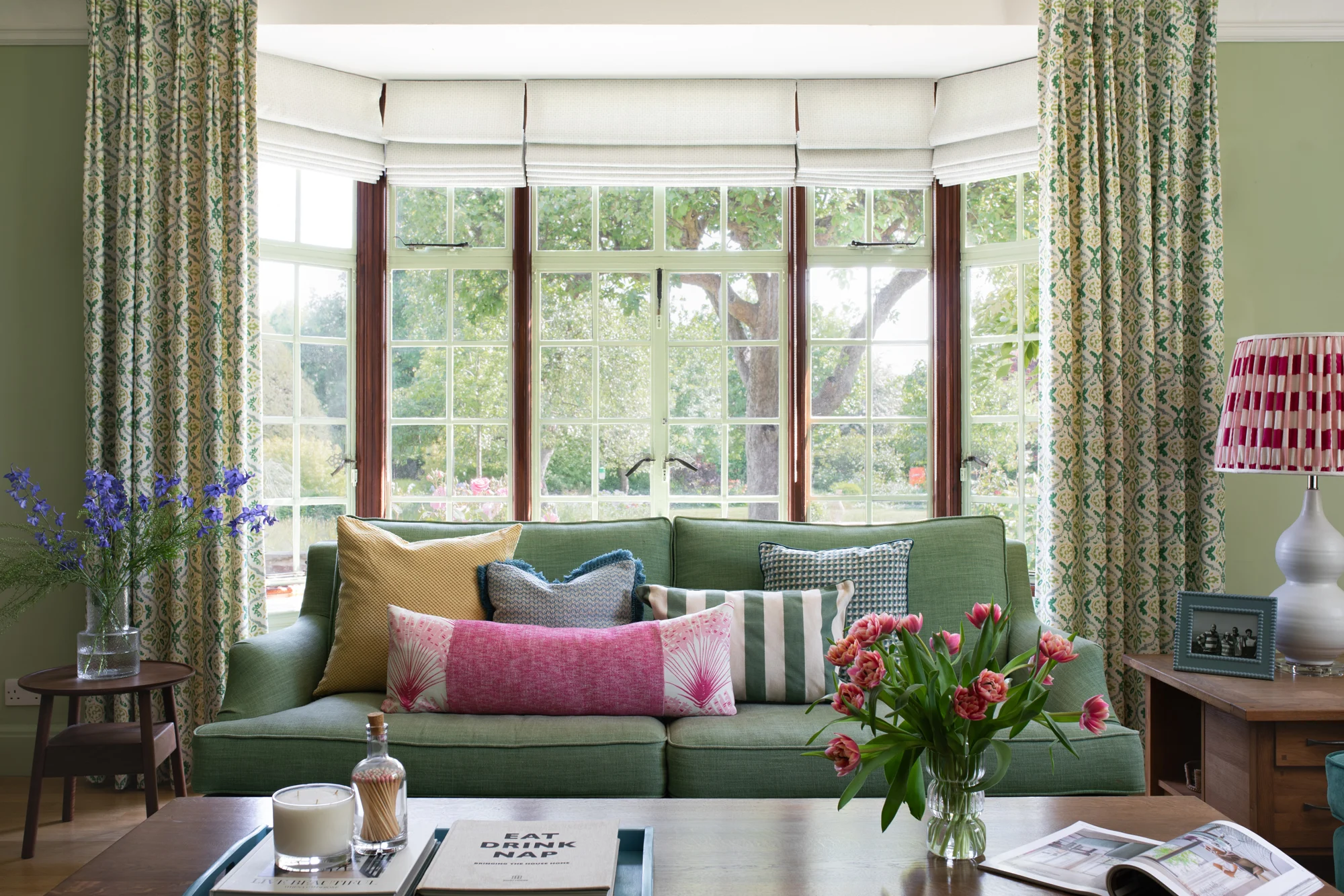 Elegant garden-inspired interior design in a light-filled living room with a green sofa, floral cushions, patterned curtains, and views of a lush garden through large bay windows.