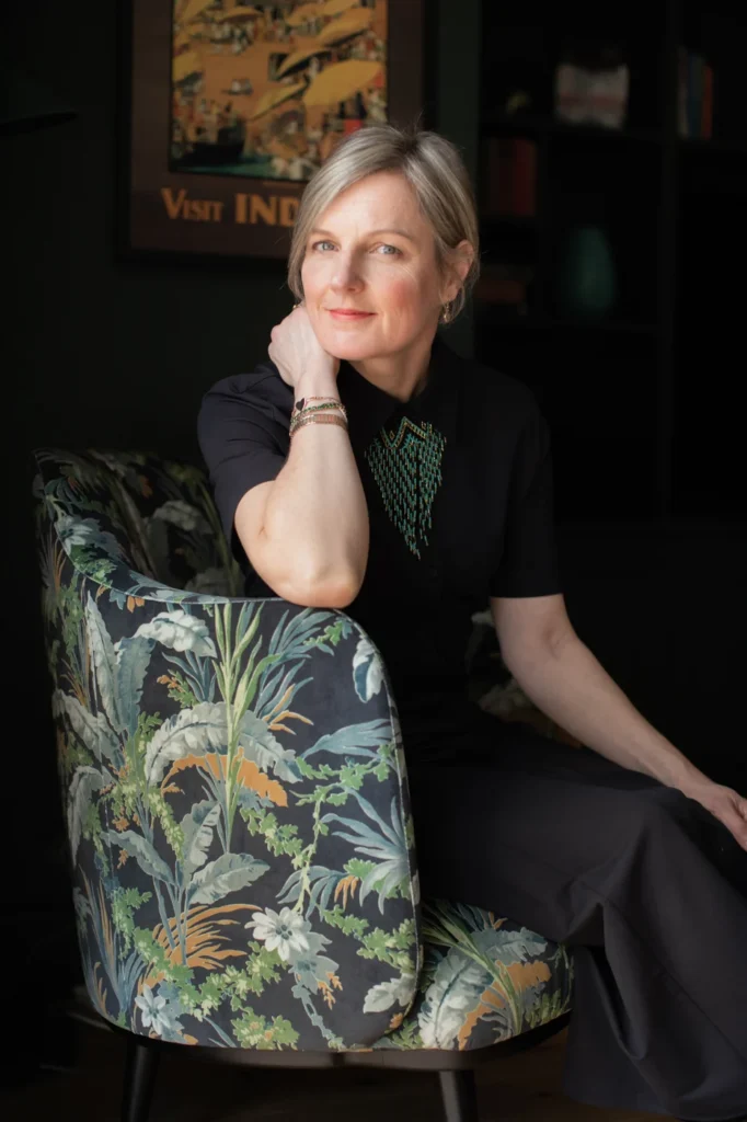 Kate Lovejoy, dressed in black with short blond hair, sitting sideways in a tub chair with colourful feature fabric