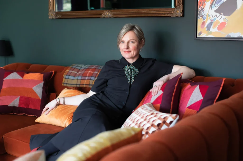 Founder and Interior Designer Kate Lovejoy in a completed home renovation feasibility project, on a rust coloured velvet sofa with patterned cushions against a teal green colour-drenched wall.