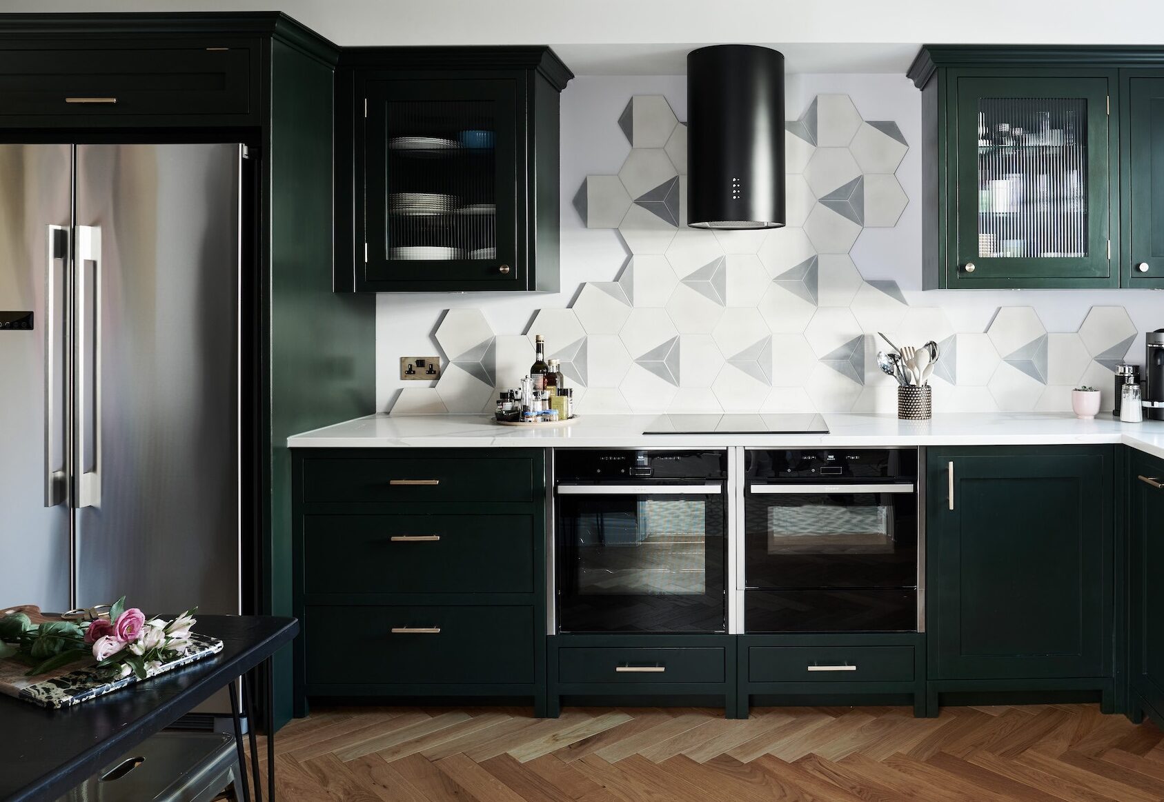 A sleek kitchen featuring dark colour palettes, with forest green cabinetry, brass handles, a white worktop, and a 3D geometric tiled backsplash set against herringbone wood flooring.