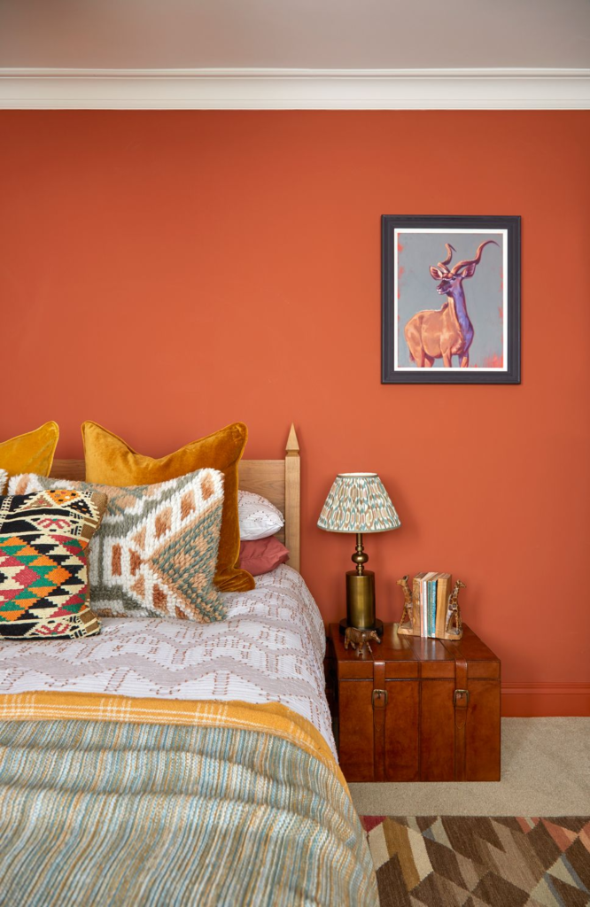 Long-term design relationship by Kate Lovejoy Interiors shows styling of a spare bedroom with an african theme, orange walls, patterned cushions and mustard yellow linens on the bed.