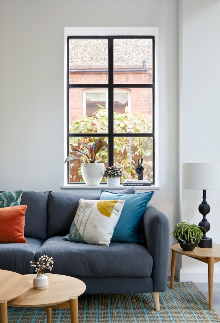 Modern design in a traditional house: open plan living area with crittall windows, a grey sofa and bright cushions, wooden coffee table and lots of plants to bring the outside in.